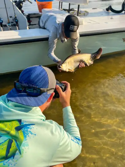 Man photographing a man hanging off the edge of a boat holding a caught fish